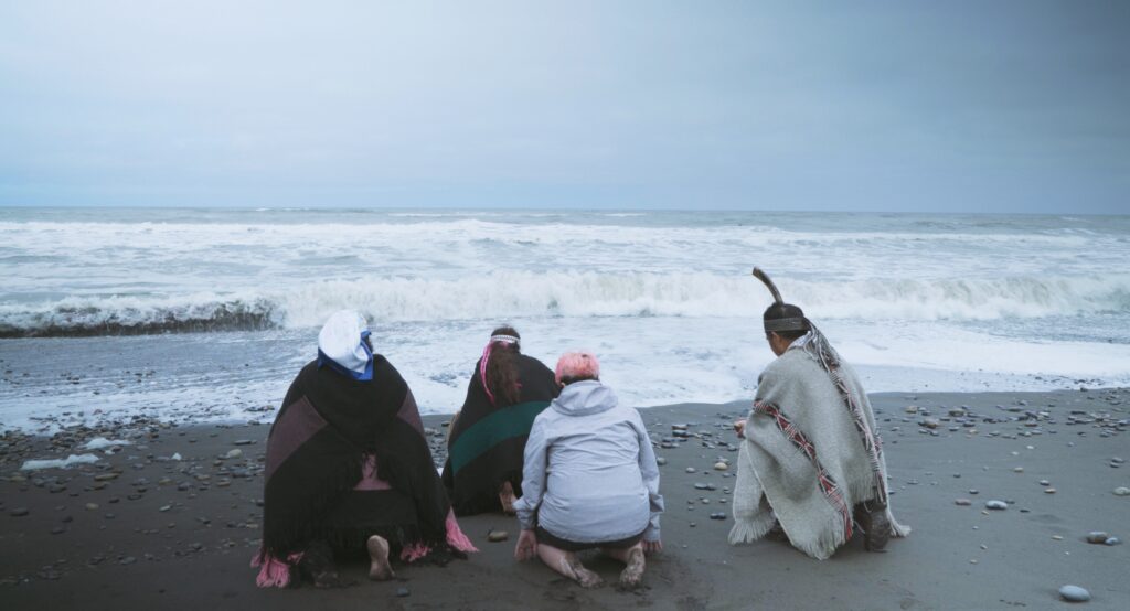 Cuatro personas, tres de ellas mapuche, hacen una ceremonia arrodilladas frente al mar.