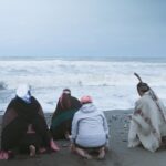 Cuatro personas, tres de ellas mapuche, hacen una ceremonia arrodilladas frente al mar.